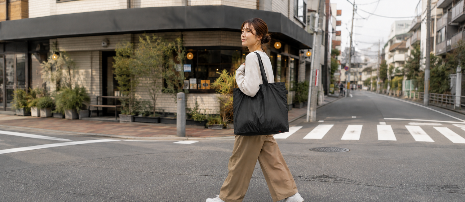 黒いトートバッグを肩掛けして街を歩く女性。日常使いにおけるpack all®と無印良品のバッグを比較するシーン